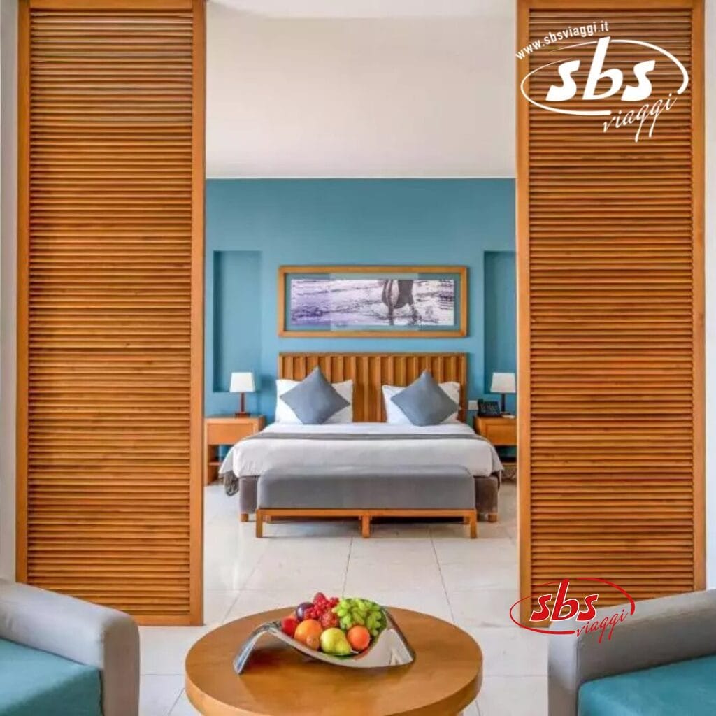 Questa accogliente camera da boutique hotel è caratterizzata da eleganti accenti blu, un grande letto invitante e due lampade dalla luce soffusa. Una ciotola di frutta fresca è appoggiata sul tavolo di legno, aggiungendo un tocco di calore. Il logo SBS Viaggi impreziosisce l'angolo in alto a destra, evidenziando il fascino del Sifawy Boutique Hotel.