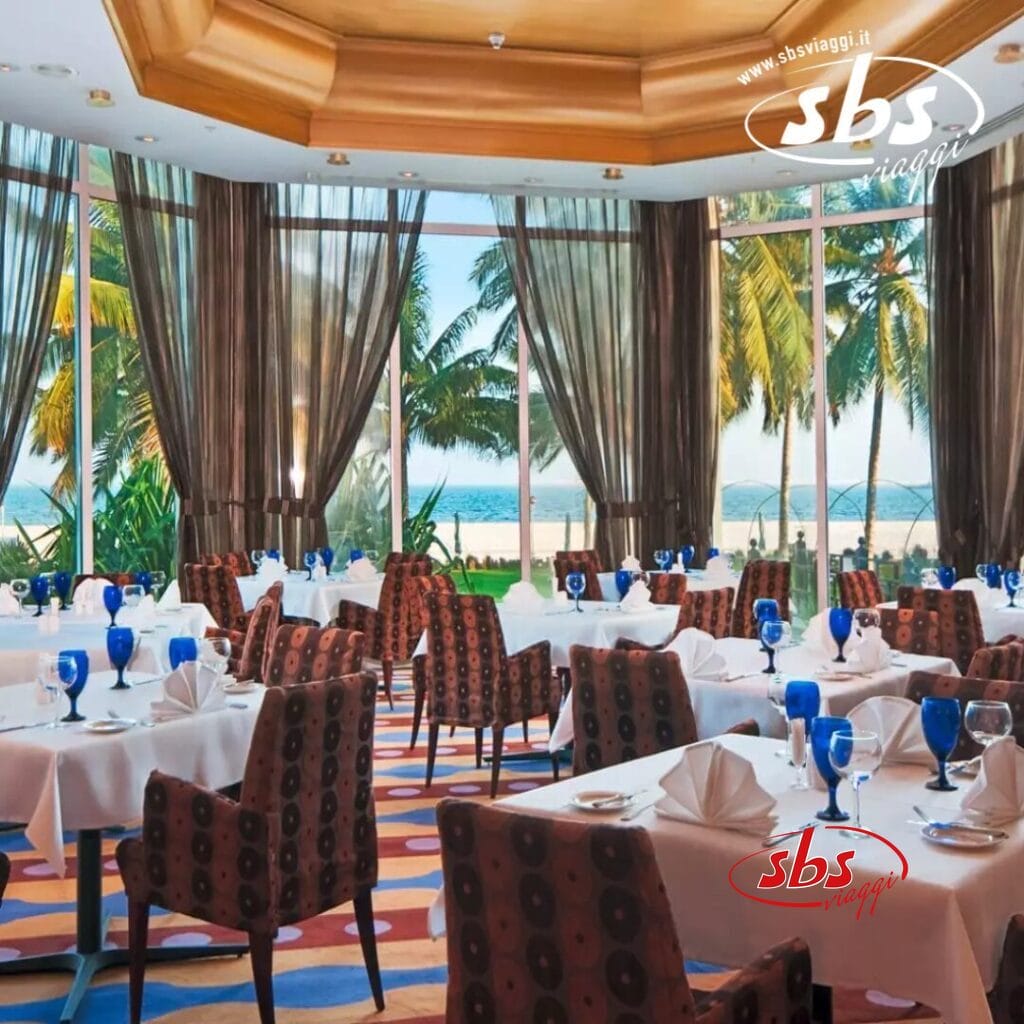 Elegante ristorante con tovaglie bianche, bicchieri blu e sedie a fantasia presso l'Hilton Resort, dotato di ampie finestre con vista sulle palme di Salalah e sull'oceano.