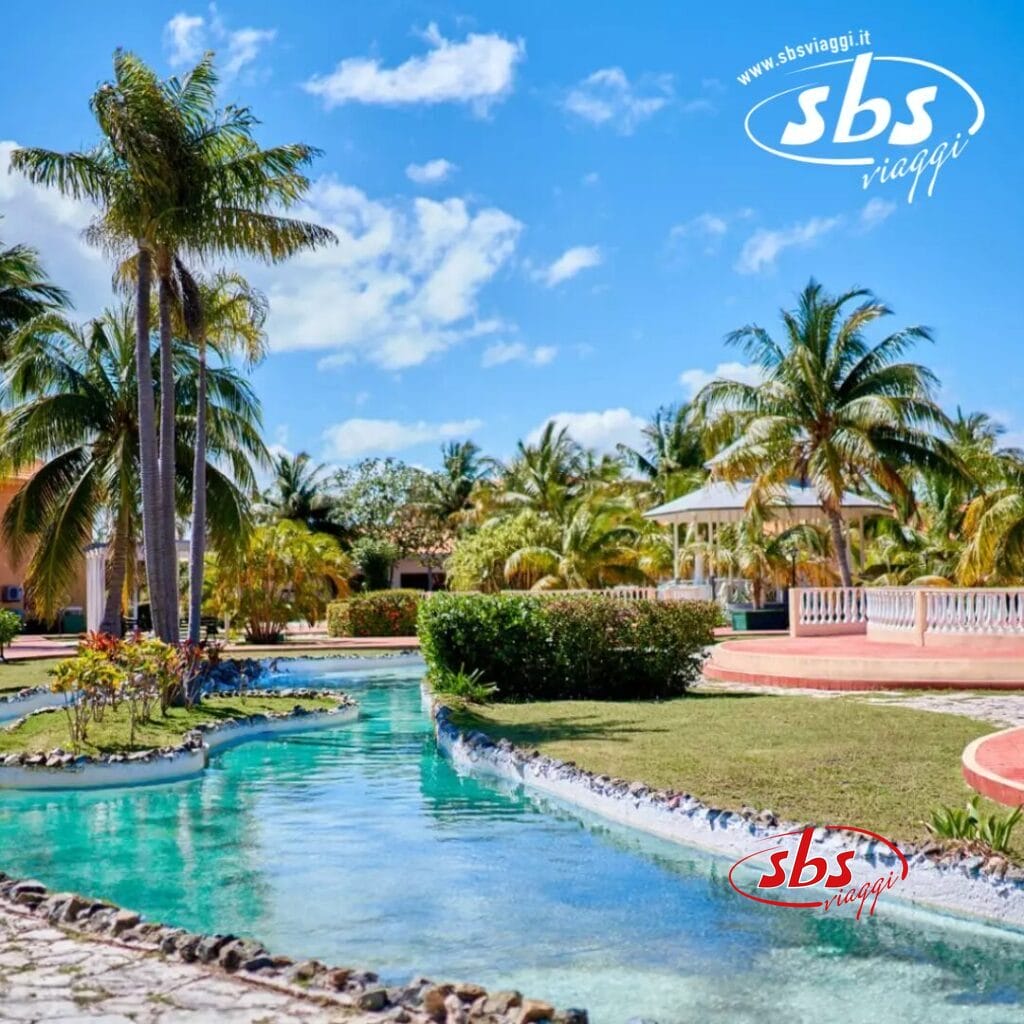 Scena di un resort tropicale con una piscina tortuosa, palme e un gazebo sotto un cielo azzurro terso. Il logo 