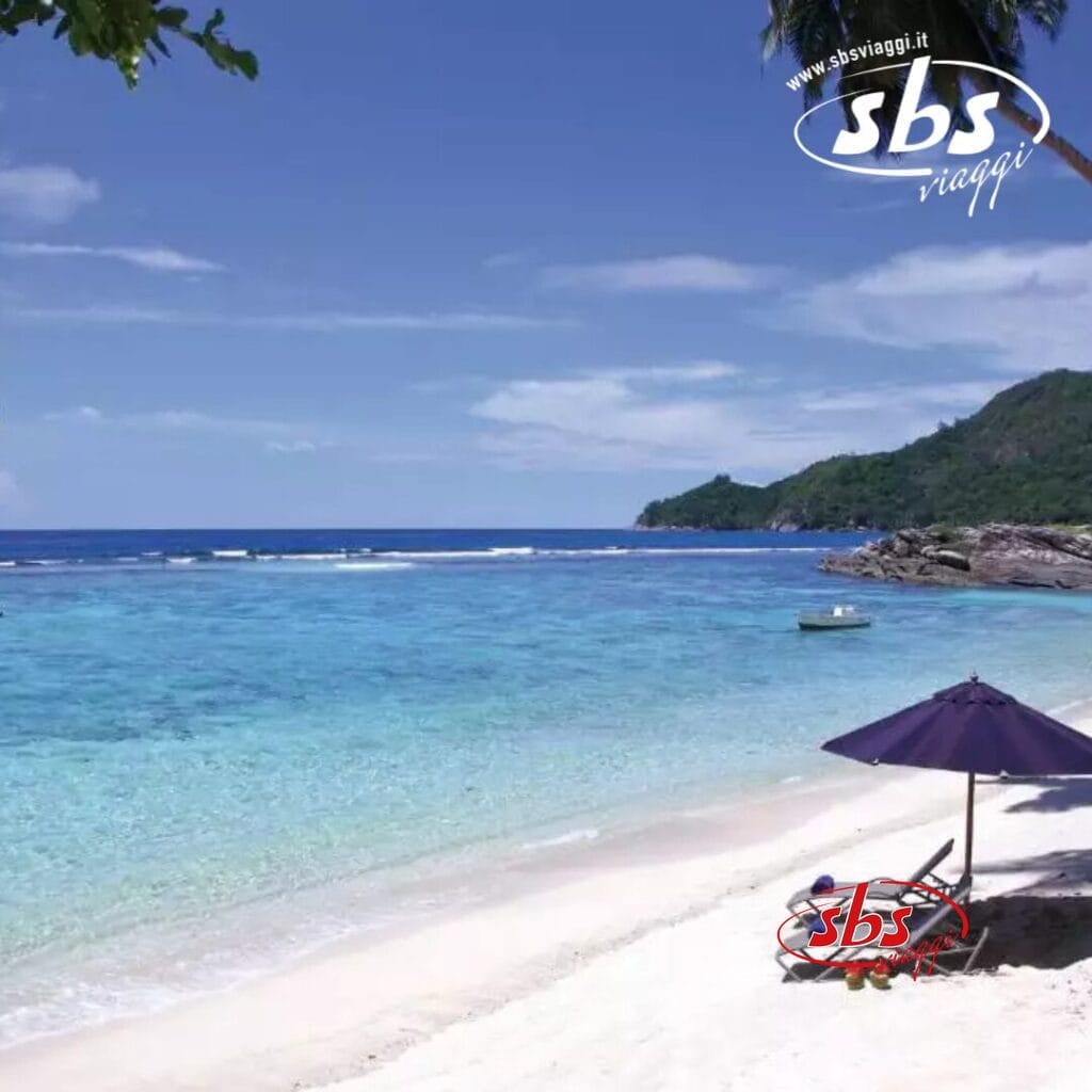 Si apre una scena di spiaggia serena con un mare calmo e cieli limpidi. Una sedia a sdraio vuota sotto un ombrellone blu invita al relax sulla sabbia, evocando una tranquillità da spa. In lontananza, le colline incorniciano lo sfondo, ricordando i tranquilli dintorni dell'Hilton Allamanda Resort.