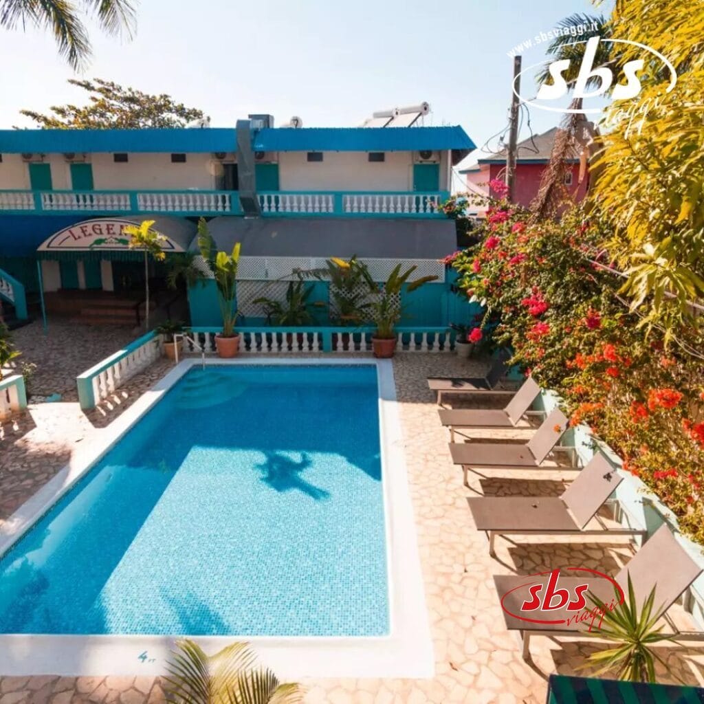 Una piscina scintilla di fronte a un edificio blu, impreziosita da sedie a sdraio su un patio in pietra e da piante tropicali che circondano l'area, creando un'invitante atmosfera di relax e tranquillità.