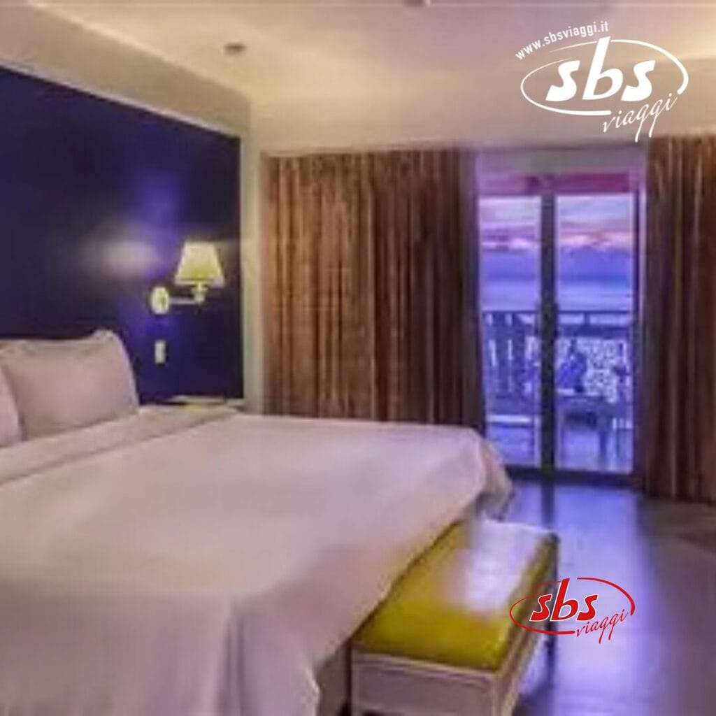 Camera d'albergo con un letto grande, lampada a parete e tende scure. Le porte del balcone rivelano una vista esterna sul Royal Decameron Montego Beach. Adatto a ospiti di età superiore ai 16 anni. Un logo SBS Viaggi è nell'angolo in alto a destra.