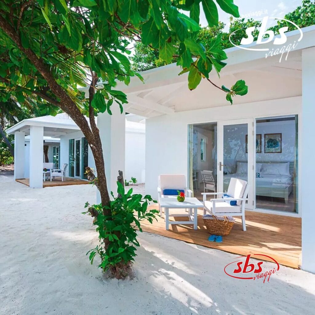 Le cabine bianche sulla spiaggia con ponti di legno, circondate da sabbia e vegetazione, creano un rifugio armonioso. Ogni cabina dispone di una pittoresca area salotto con sedie e un tavolo, perfetta per il relax. La scena sembra che potrebbe essere catturata da una bozza automatica, preservando il suo fascino sereno senza sforzo.