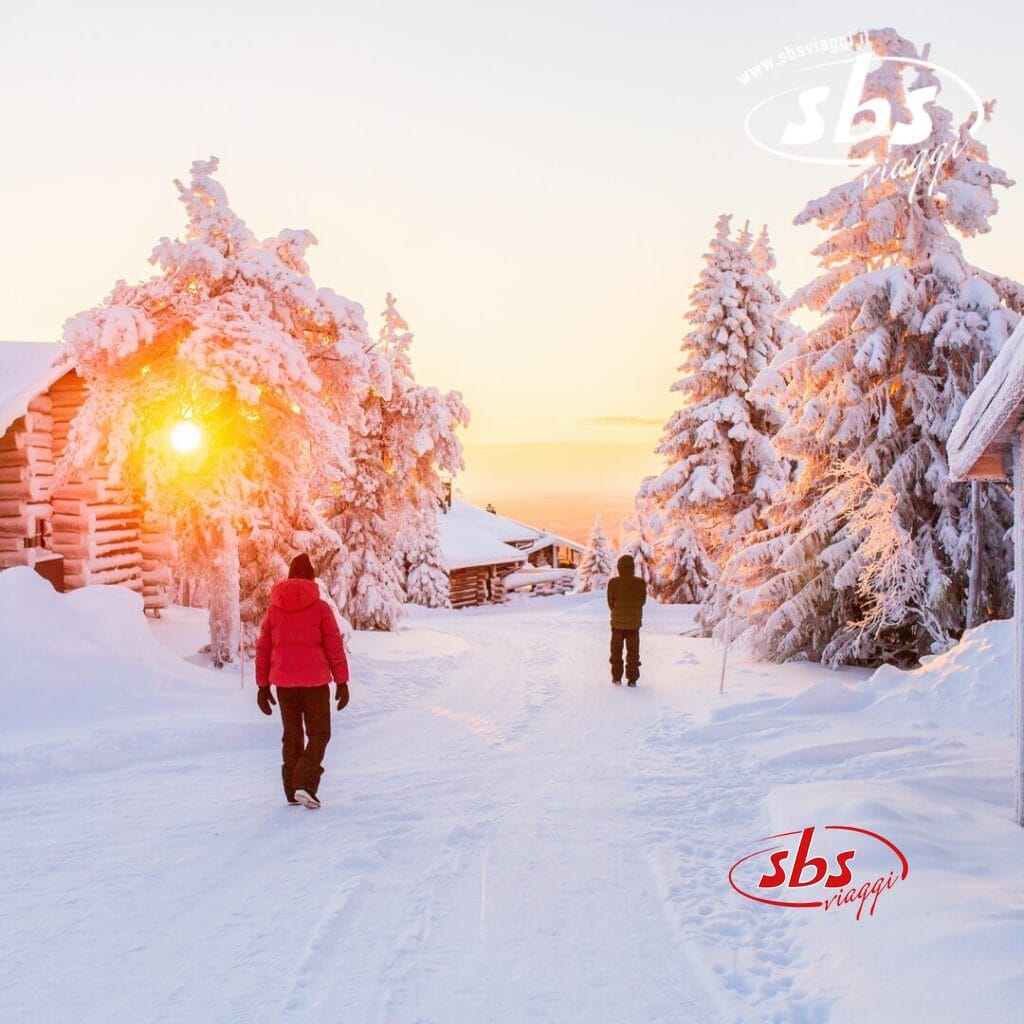 Due persone passeggiano lungo un sentiero innevato a Rovaniemi, circondate da alberi innevati e accoglienti baite, mentre il sole tramonta come una favola invernale.