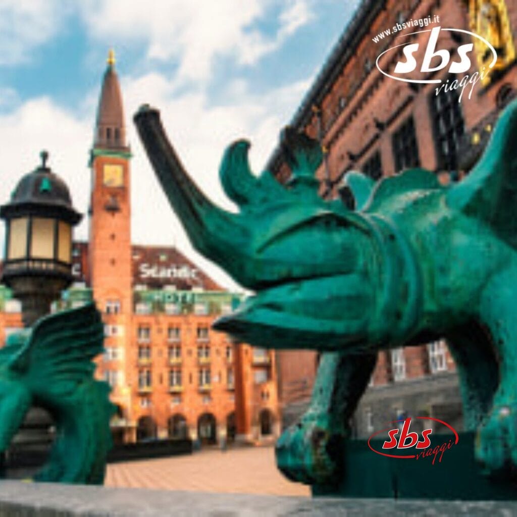 Primo piano di una scultura di rinoceronte verde di fronte a uno storico edificio in mattoni con torre e orologio, che ricorda il fascino architettonico di Copenaghen. Il logo di SBS Viaggi è nell'angolo in alto a destra.