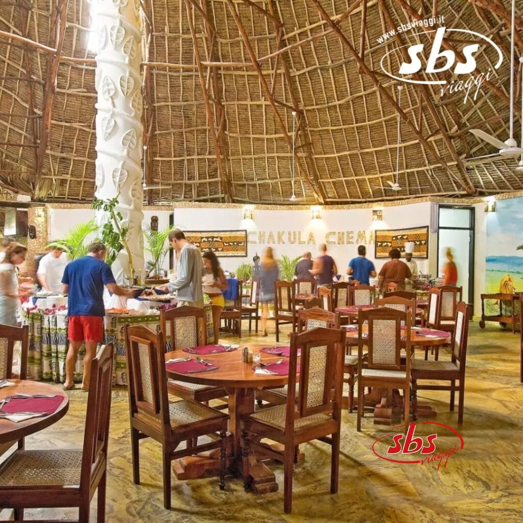 Un ristorante interno a tema tropicale con tavoli e sedie in legno offre un'atmosfera vivace. Gli ospiti sono in piedi al buffet vicino a un pilastro decorativo sotto un tetto di paglia, creando una scena vibrante come Bozza automatica in azione.