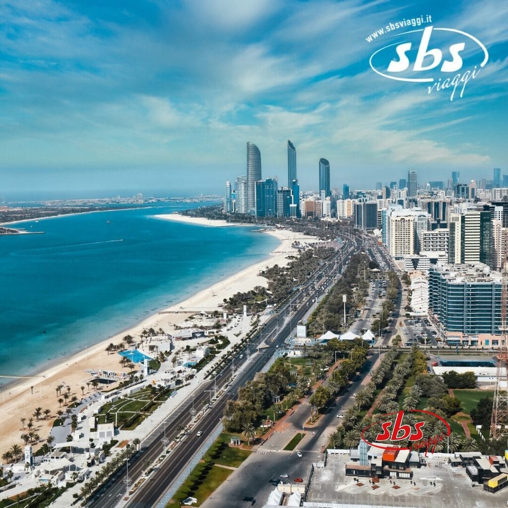 Vista aerea di una città costiera degli Emirati Arabi Uniti con grattacieli, spiaggia sabbiosa e strada fiancheggiata da palme sotto un cielo nuvoloso, che mostra uno scenario degno di un Grand Tour.