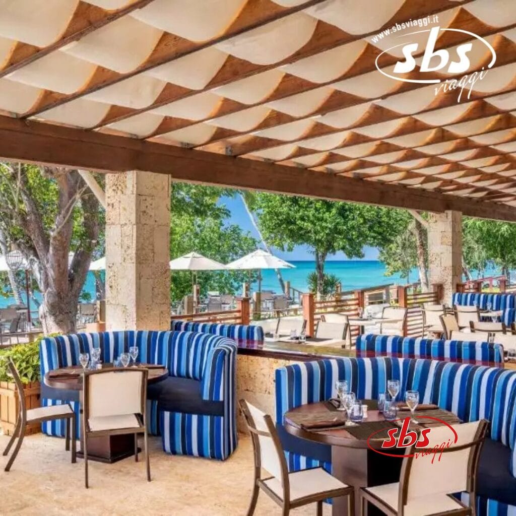 Situato a La Romana, questo ristorante all'aperto è caratterizzato da sedute blu a strisce con tavoli e sedie. Affacciato sulla spiaggia con alberi, ombrelloni bianchi e vista sul mare, fa parte di un affascinante resort per famiglie. Un tetto decorato con motivi geometrici completa l'ambiente invitante.