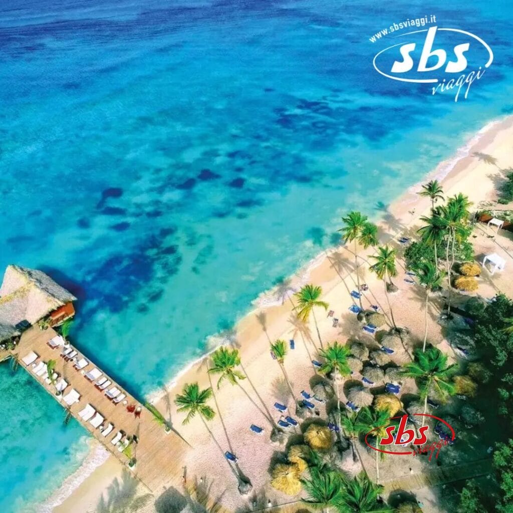 Vista aerea della spiaggia tropicale del Veraclub, con palme, un molo di legno e ombrelloni di paglia, bagnati da acque cristalline.