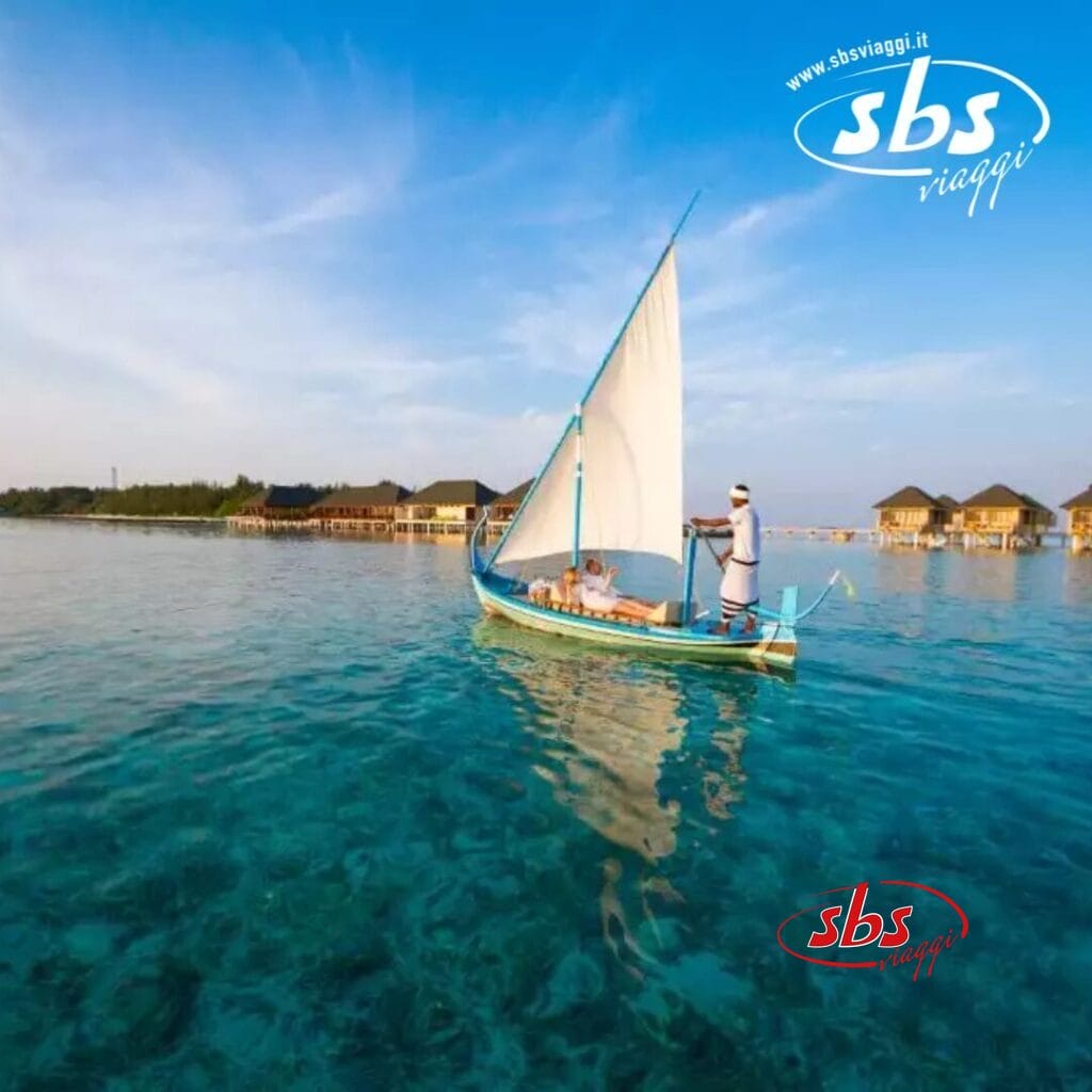 Una persona naviga su una piccola barca con una vela bianca su acque azzurre e limpide, godendosi il calore di un'isola estiva vicino ai bungalow sull'acqua sotto un cielo azzurro brillante.