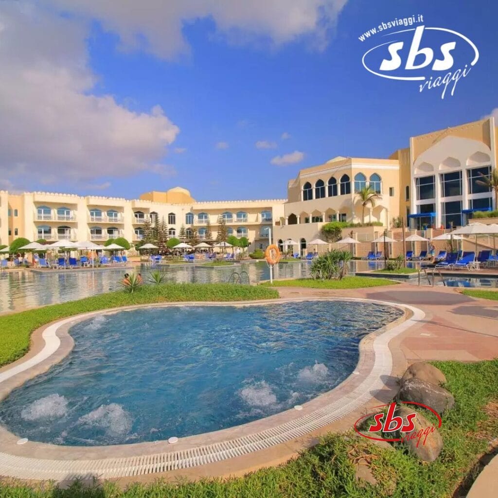 La piscina all'aperto e l'area prendisole, perfetta per rilassarsi, si trovano accanto al grande edificio dell'hotel, con balconi e sotto un cielo azzurro e terso.