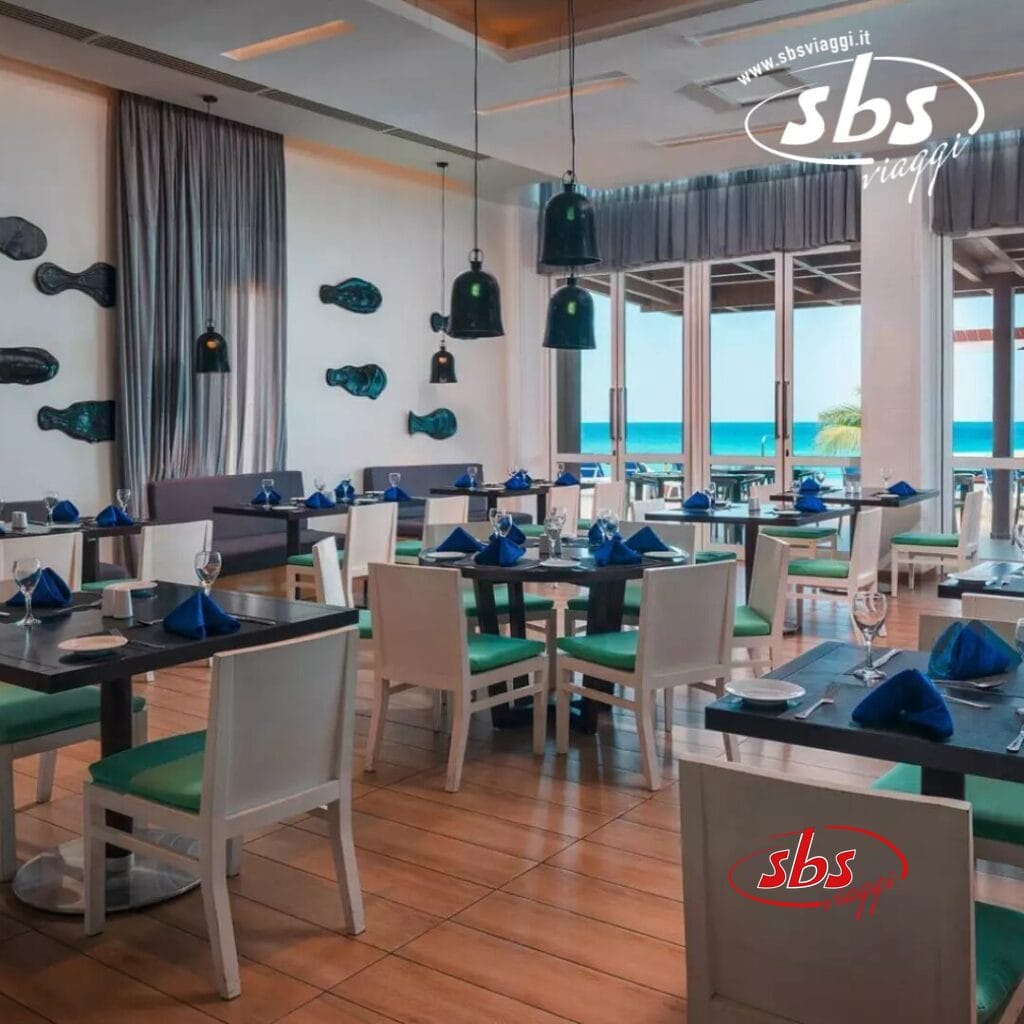 Gli interni moderni del ristorante sono caratterizzati da tavoli apparecchiati con cura, decorati con tovaglioli blu e grandi finestre che regalano una vista sull'oceano, mentre le luci del soffitto aggiungono un tocco di eleganza, creando un'atmosfera che ricorda Bozza Automatica.