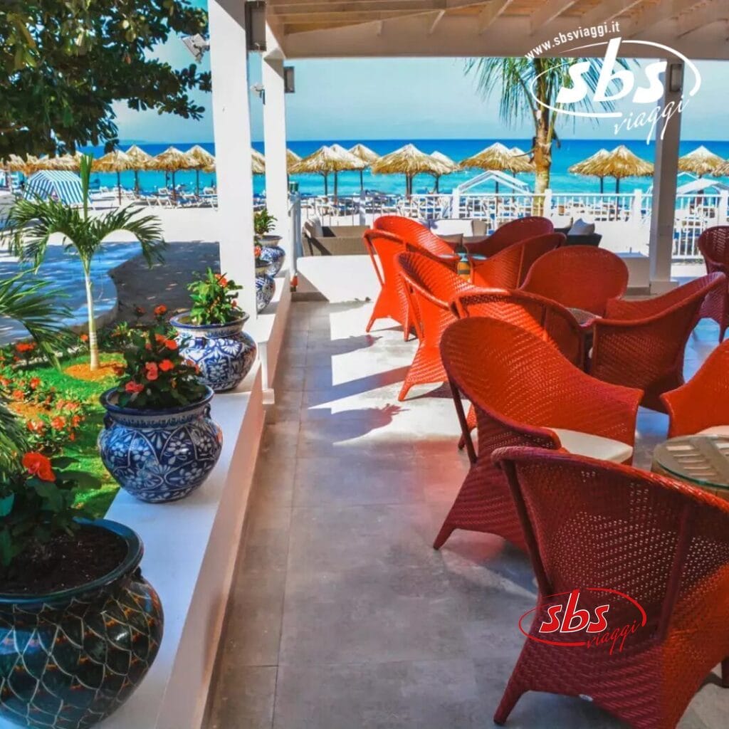 Patio fronte mare con sedie rosse, piante in vaso e vista sull'oceano. Bozza automatica cattura file di ombrelloni da spiaggia visibili sullo sfondo sotto un cielo azzurro terso.