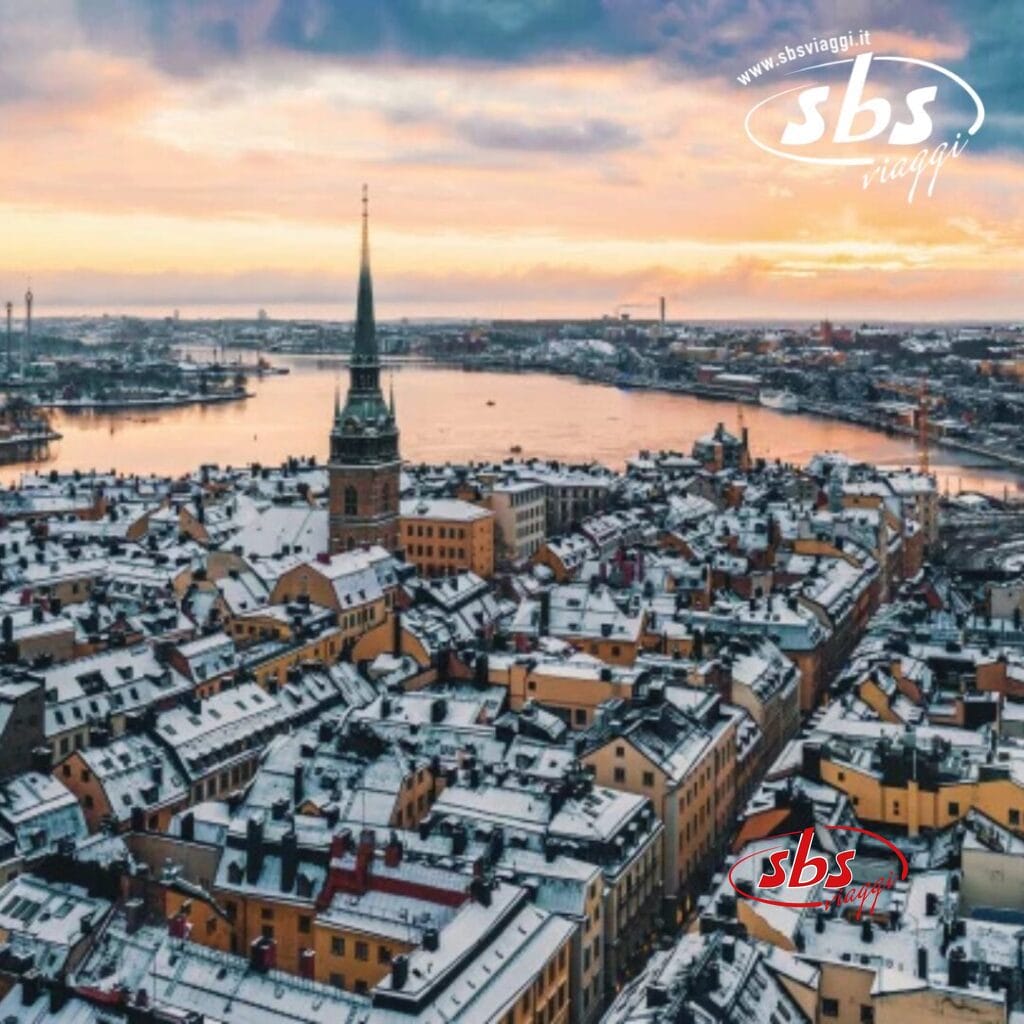 Vista aerea di un paesaggio urbano innevato con un importante campanile di chiesa, circondato dall'acqua e immerso in un tramonto dorato, che cattura la magia delle capitali del Nord. Il logo di 