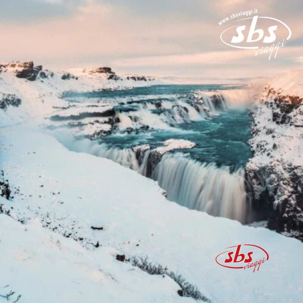 Un paesaggio innevato in Islanda presenta una grande cascata che si riversa su più livelli, circondata da terreno roccioso. Il cielo è coperto con accenni di aurore boreali e un logo con il testo 