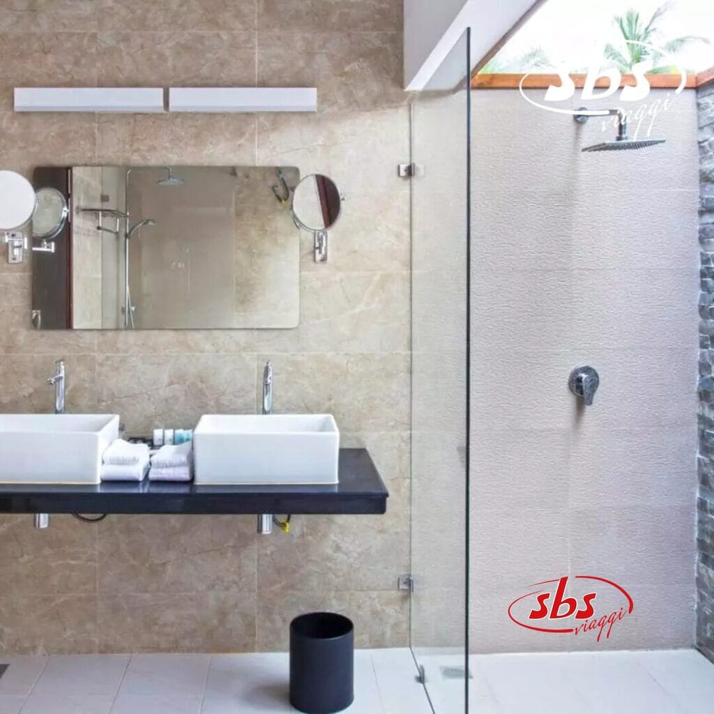 Il moderno bagno del Pearl Sands è dotato di doppi lavandini, un grande specchio e una cabina doccia con divisorio in vetro. Le pareti sono decorate con piastrelle beige, completate da una piccola finestra sopra la zona doccia. È un'oasi di tranquillità che riflette l'eleganza di un boutique hotel.