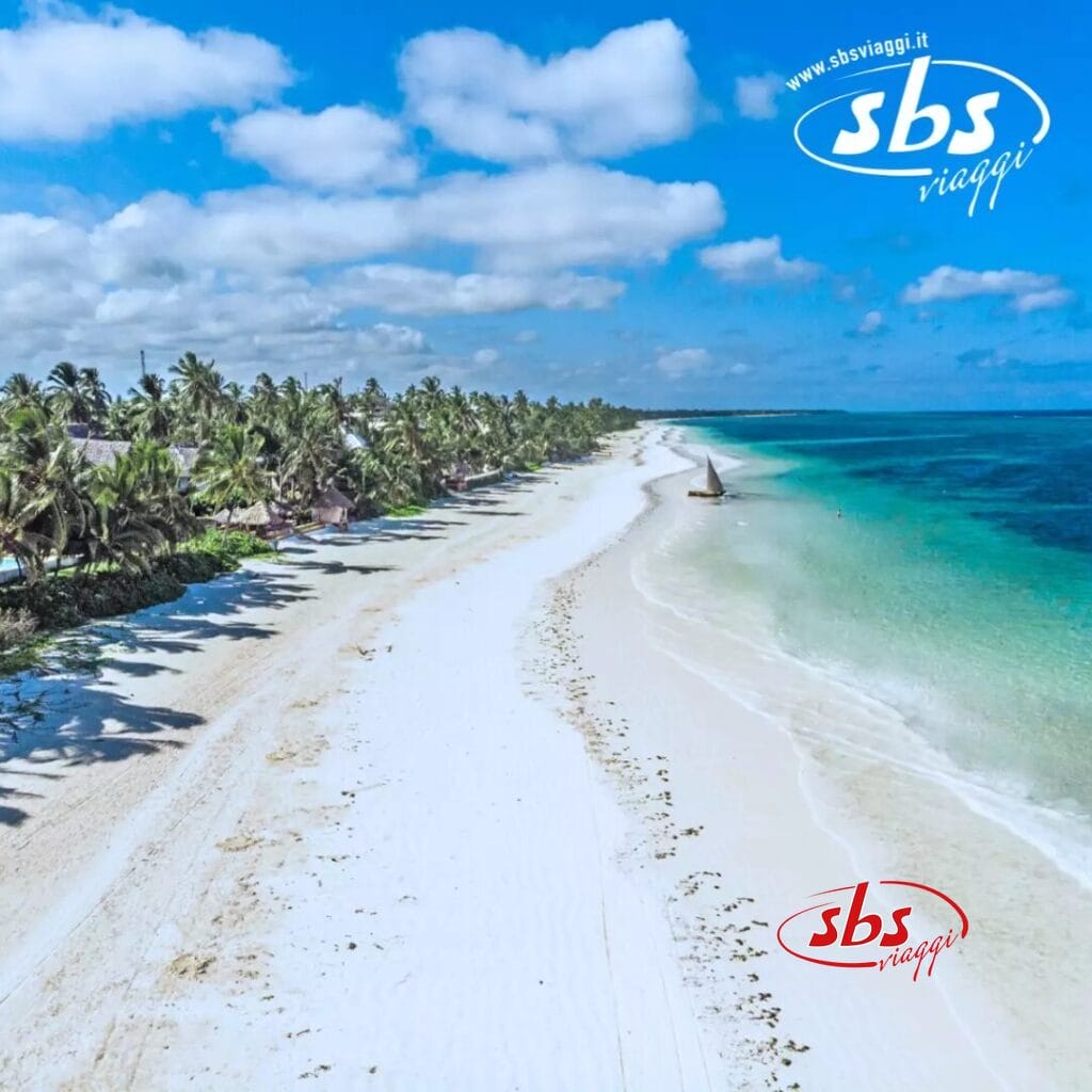 Vista aerea di una spiaggia tropicale vicino al Sbh Kilindini Resort, con sabbia bianca, palme e acqua turchese. Una piccola barca a vela scivola vicino alla riva sotto un cielo parzialmente nuvoloso.