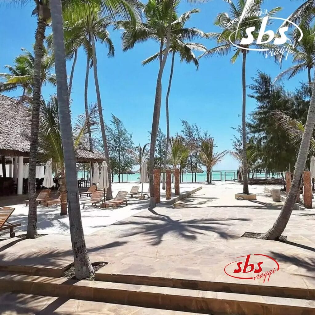 Una scena di spiaggia tropicale a Zanzibar presenta palme, lettini prendisole e un cielo azzurro e limpido. Una struttura con tetto di paglia è sulla sinistra e l'oceano luccica sullo sfondo, esaltando questo paradiso del resort.