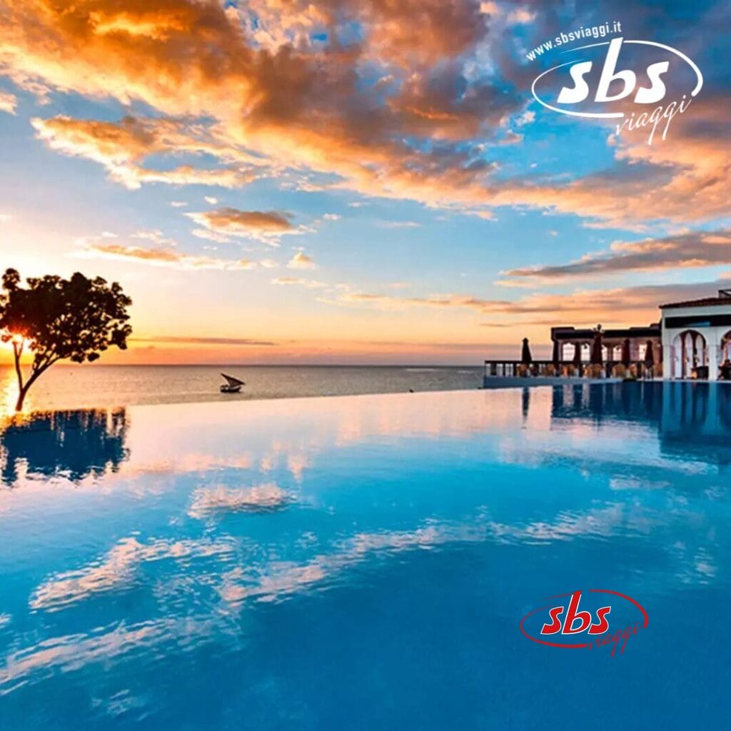 Piscina a sfioro al Riu Palace Zanzibar con vista sull'oceano, con cielo al tramonto, sagome di alberi e area lounge in lontananza. Logo SBS travel nell'angolo. Vivi l'atmosfera da hotel di lusso per eccellenza in questo incontaminato resort di Zanzibar.