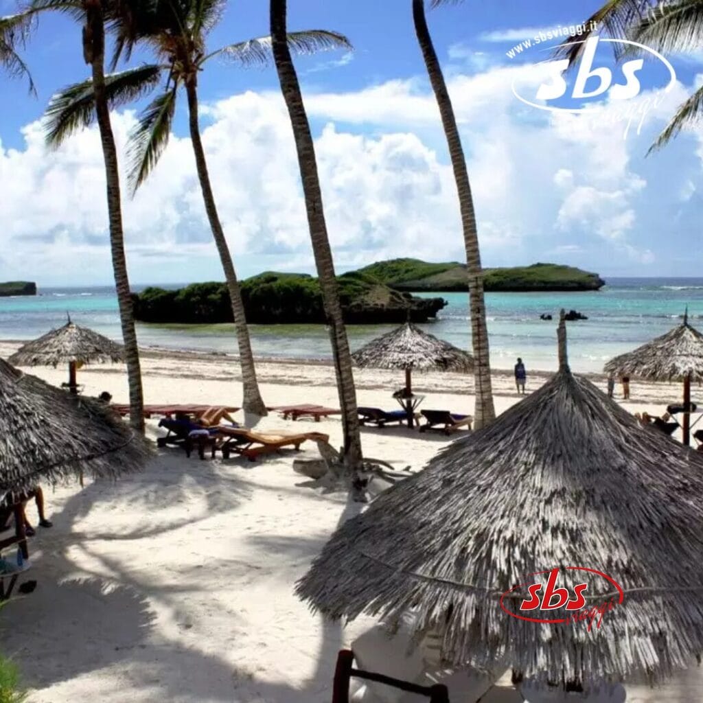 Scena di spiaggia tropicale a Crystal Bay, con palme, ombrelloni di paglia e le comode sedie a sdraio del Veraclub di fronte all'oceano. Una piccola isola è visibile in lontananza sotto un cielo parzialmente nuvoloso.