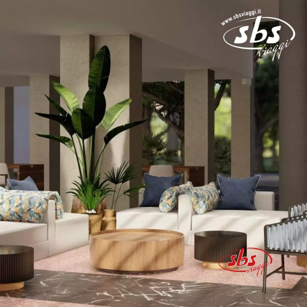 Una moderna area lounge al Coral Costa Caribe presenta divani bianchi, cuscini fantasia, una grande pianta e tavoli rotondi in legno su un pavimento in marmo. Il logo "sbs viaggi" è elegantemente esposto in alto a destra.