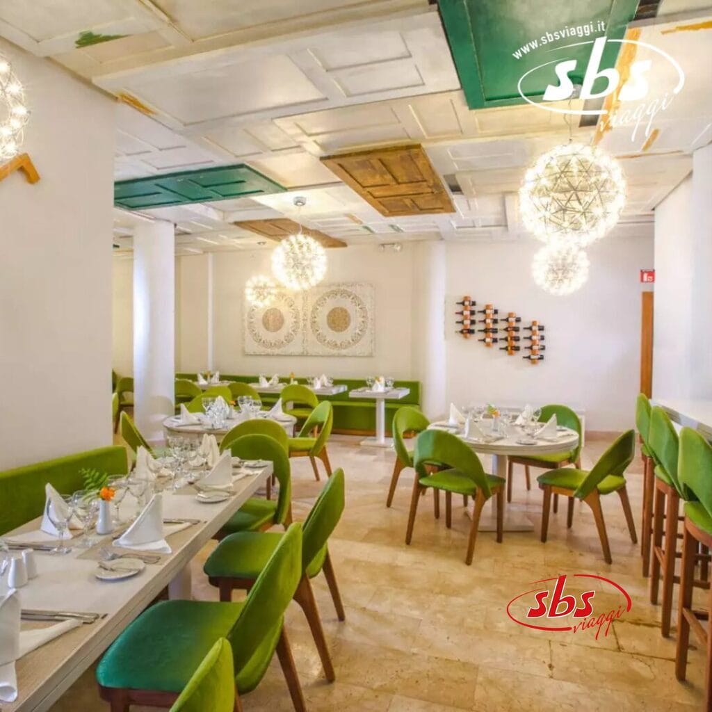 Un moderno ristorante interno con sedie Bozza automatica-verde e tavoli bianchi apparecchiati per la cena, il tutto sotto lampadari decorativi. Le pareti vantano opere d'arte, completate da pannelli sul soffitto in un armonioso mix di toni verdi e marroni.