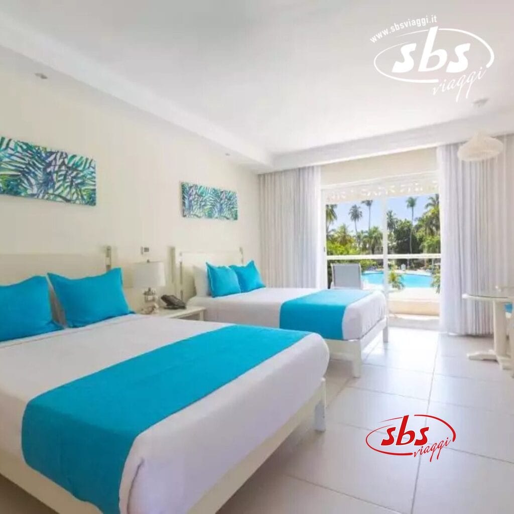 Camera d'albergo con due letti con accenti turchesi, un tavolino e una vista sulla piscina e sul giardino tropicale attraverso grandi finestre nel nostro resort sulla spiaggia di Punta Cana. Godetevi l'accesso alla nostra lussuosa spa per un soggiorno davvero rilassante.