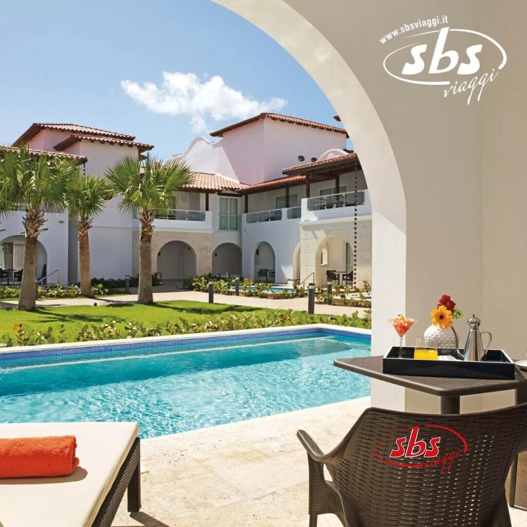 Vista a bordo piscina di un resort in vero stile Seaclub, con edifici bianchi, palme e uno splendido cielo azzurro. Un tavolo con bevande e una sedia sono in primo piano. Questa fuga serena cattura l'essenza del Dreams Dominicus nella pittoresca La Romana.