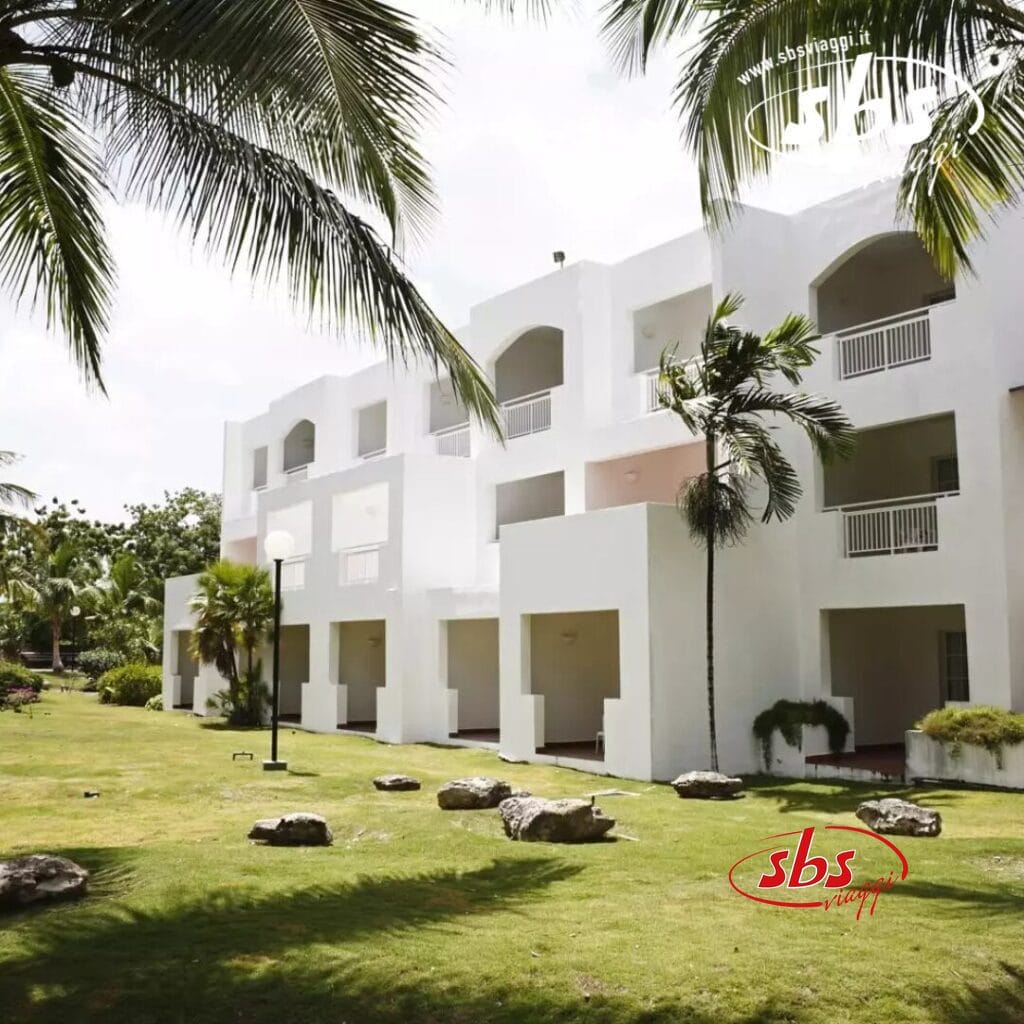 Immerso tra rigogliose palme tropicali, il moderno edificio bianco del Veraclub Dominicus è dotato di eleganti balconi che si affacciano su un prato erboso sotto un cielo nuvoloso.