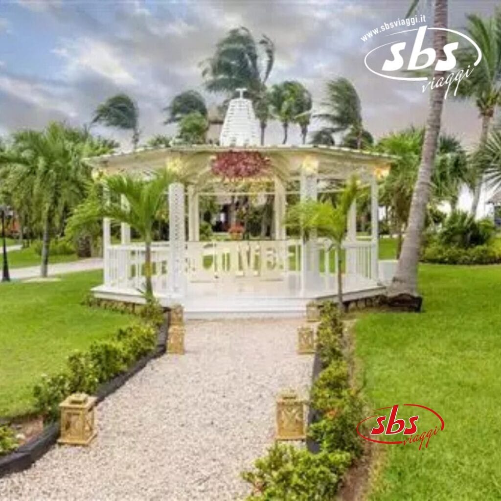 Un gazebo bianco, simile a una bozza automatica nel design della natura, riposa serenamente tra palme e vegetazione lussureggiante, con un sentiero di ghiaia che conduce con grazia ad esso sotto il morbido velo di un cielo nuvoloso.