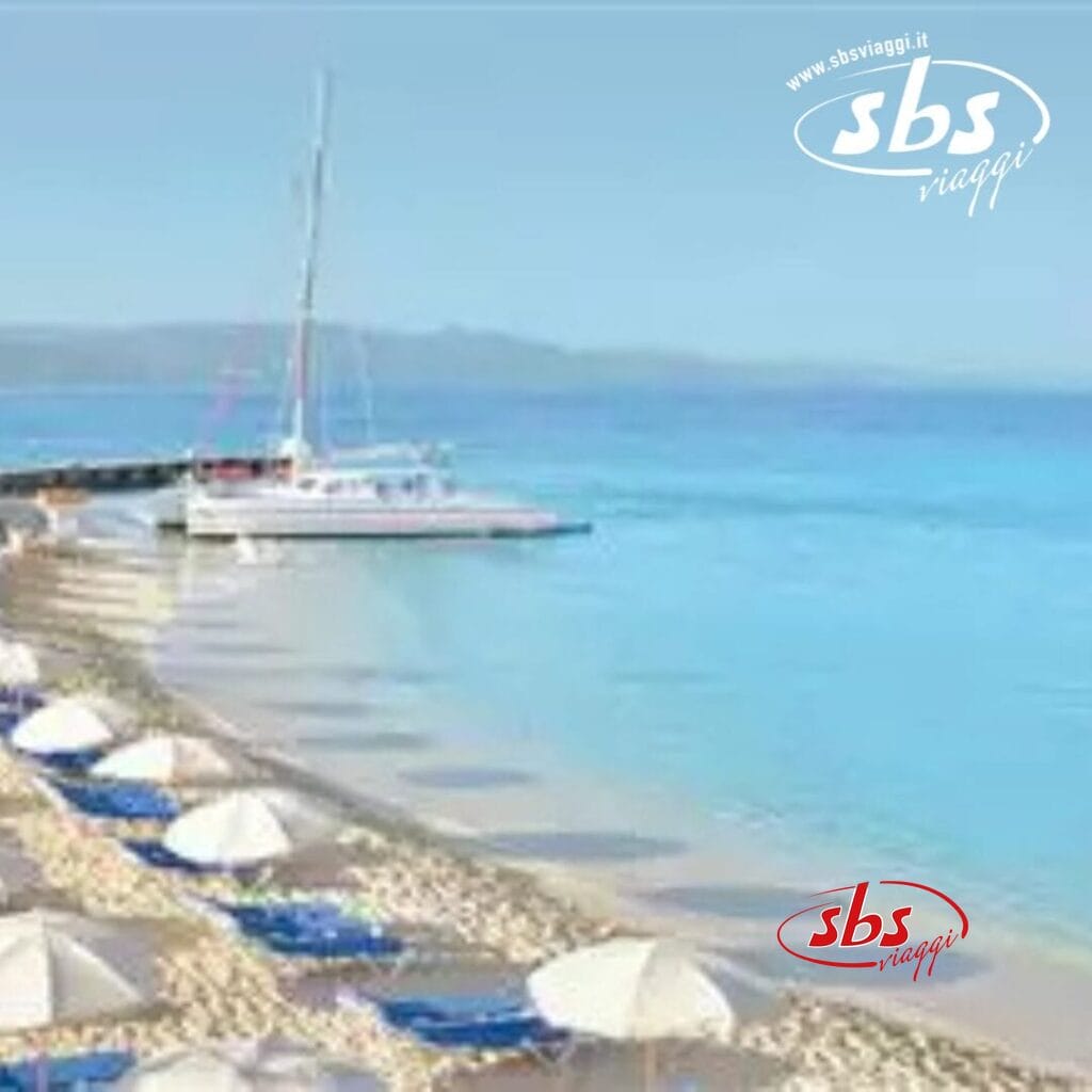 Un catamarano riposa sulle rive sabbiose del Royal Decameron Montego Beach, circondato da ombrelloni e sedie a sdraio blu. L'acqua cristallina luccica sotto un cielo azzurro. Perfetto per gli ospiti di età superiore ai 16 anni in cerca di tranquillità. Logo nell'angolo in alto a destra.