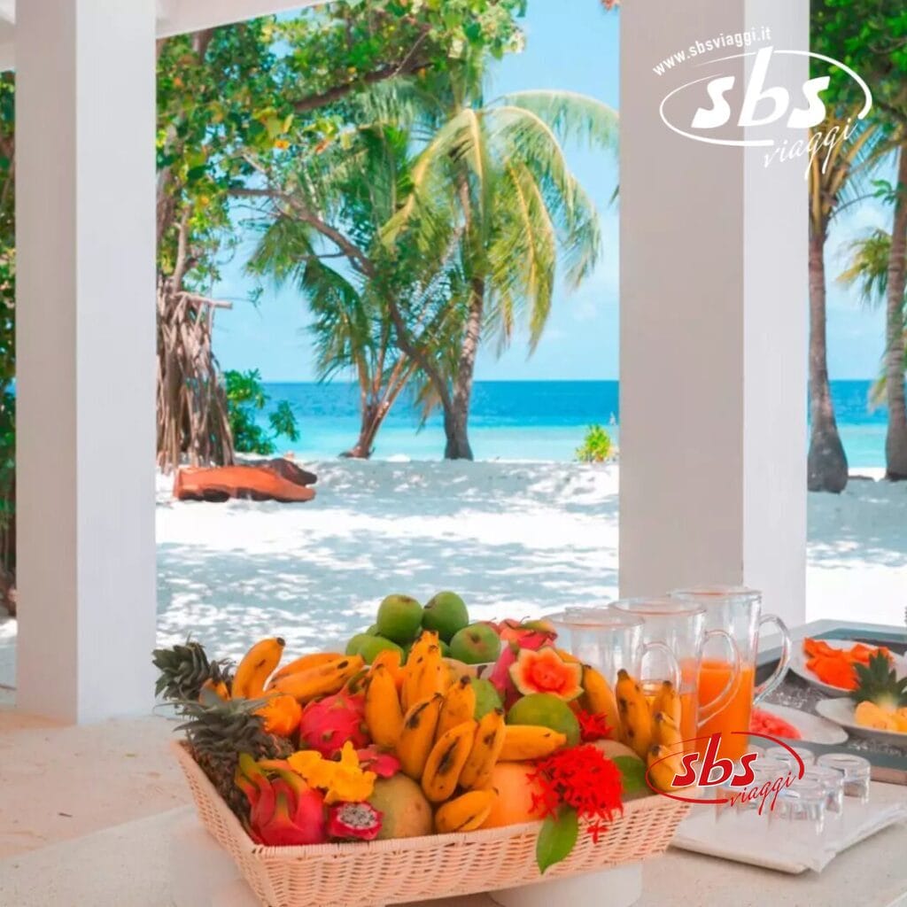 Un cesto di frutta tropicale e una brocca di succo sono appoggiati su un tavolo con vista sulla spiaggia, sulle palme ondeggianti e sul rumore dell'oceano blu.