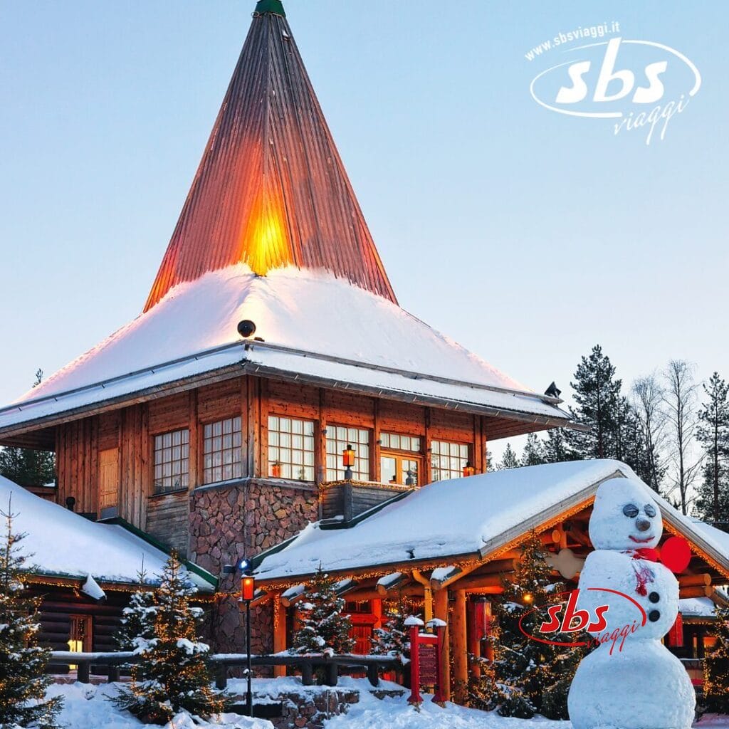 Edificio in legno ricoperto di neve con un alto tetto triangolare, incastonato tra i pini in Lapponia finlandese. Un allegro pupazzo di neve si erge fiero di fronte. Logo 