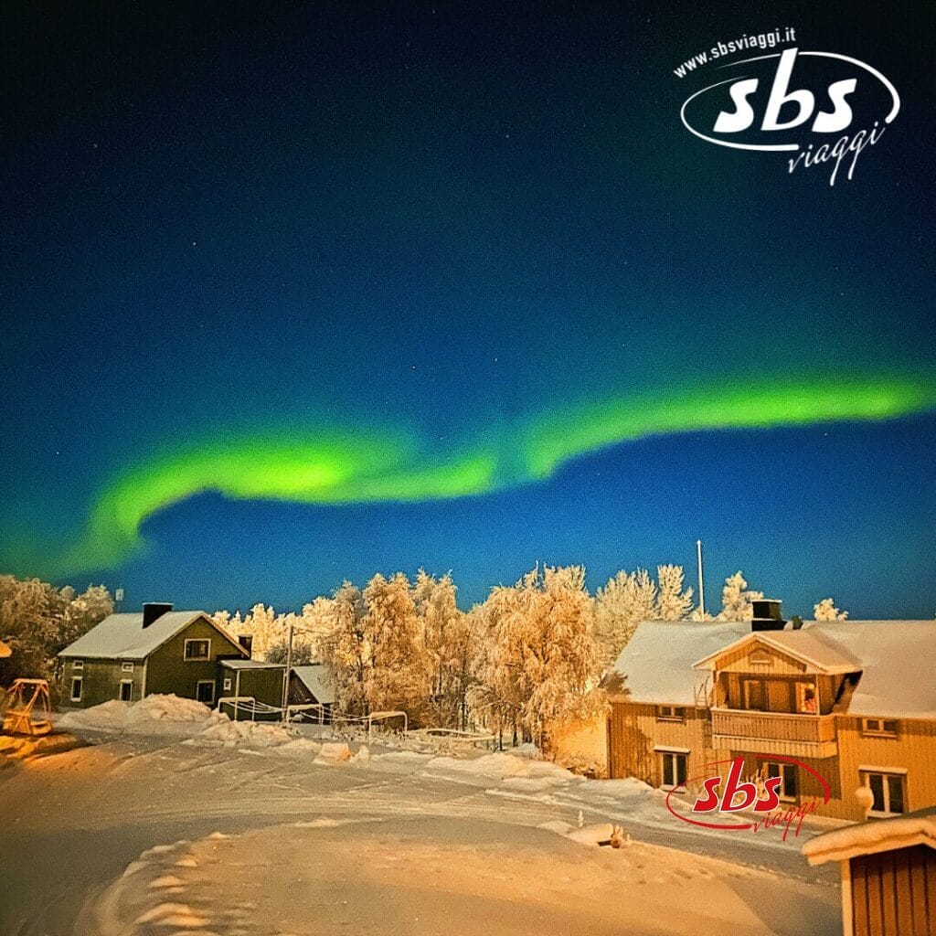 L'aurora boreale illumina il cielo notturno sopra le case e gli alberi innevati, trasformando il paesaggio di Rovaniemi in una favola invernale.