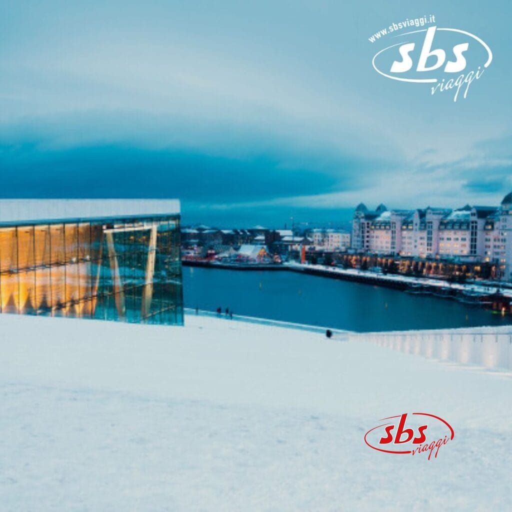 In un'affascinante scena invernale, un moderno edificio in vetro si erge accanto a fiordi sereni, mentre la sponda opposta rivela un'architettura tradizionale. Il cielo coperto aggiunge fascino. Il logo recita 