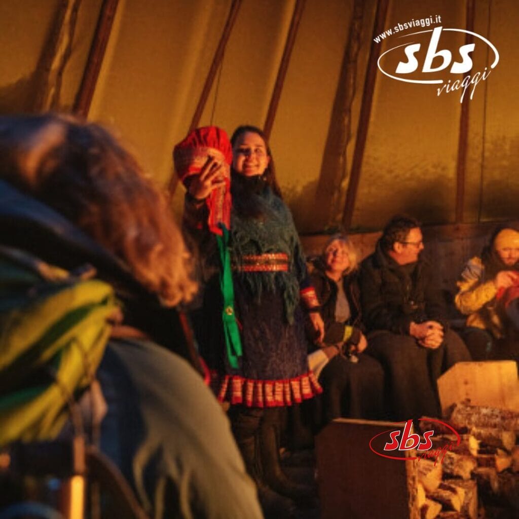 Un gruppo di persone siede all'interno di una tenda, riunite attorno a un fuoco. Una persona è in piedi con un oggetto in mano, mentre gli altri sono seduti e guardano. Fuori, la mistica aurora boreale danza sul paesaggio artico vicino a Tromso. Un logo con la scritta 