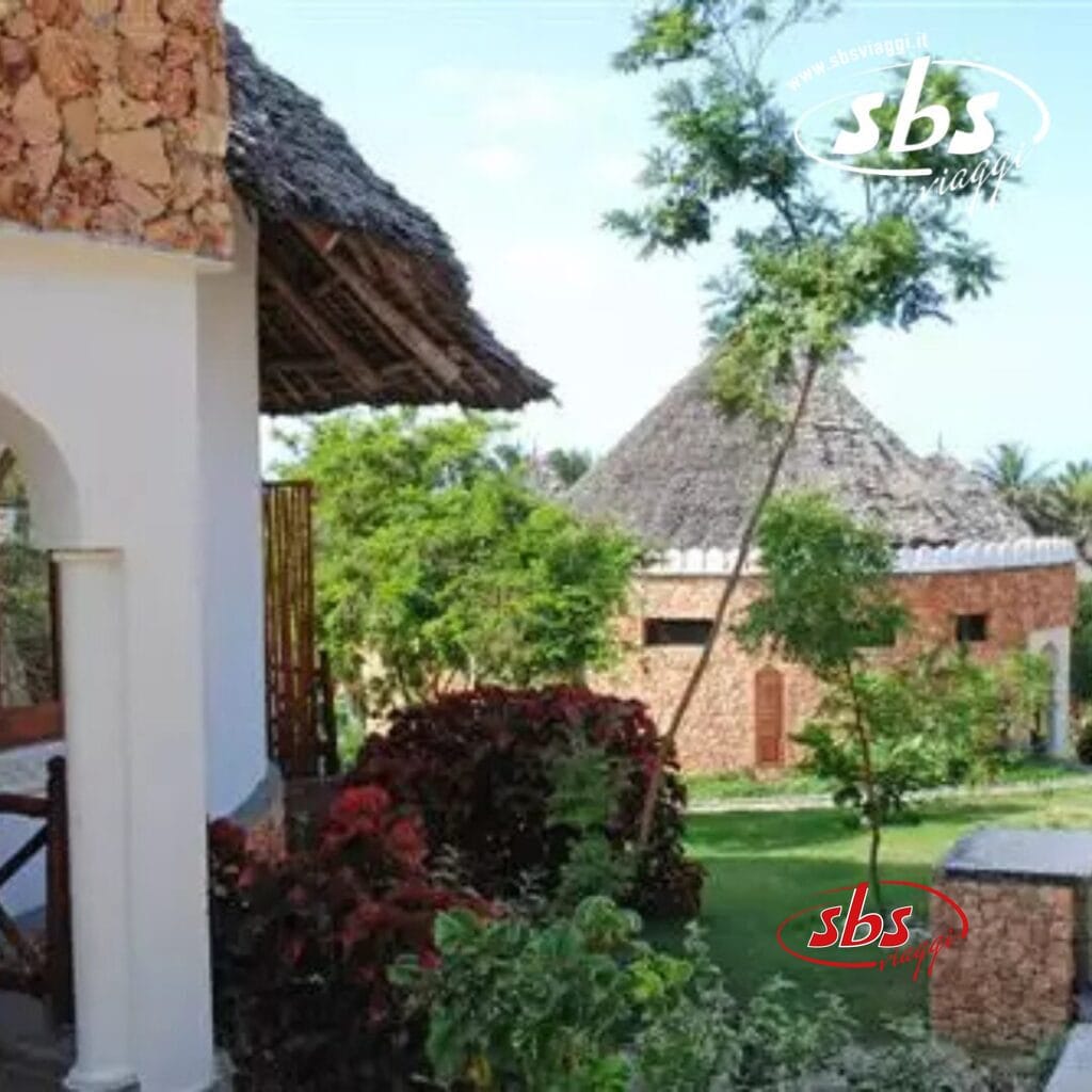 Vista sul giardino di edifici rustici con tetti di paglia, circondati da una vegetazione lussureggiante e da alberi sotto un cielo azzurro e terso, che offrono uno scenario così armonioso da sembrare una bozza automatica creata dalla natura stessa.