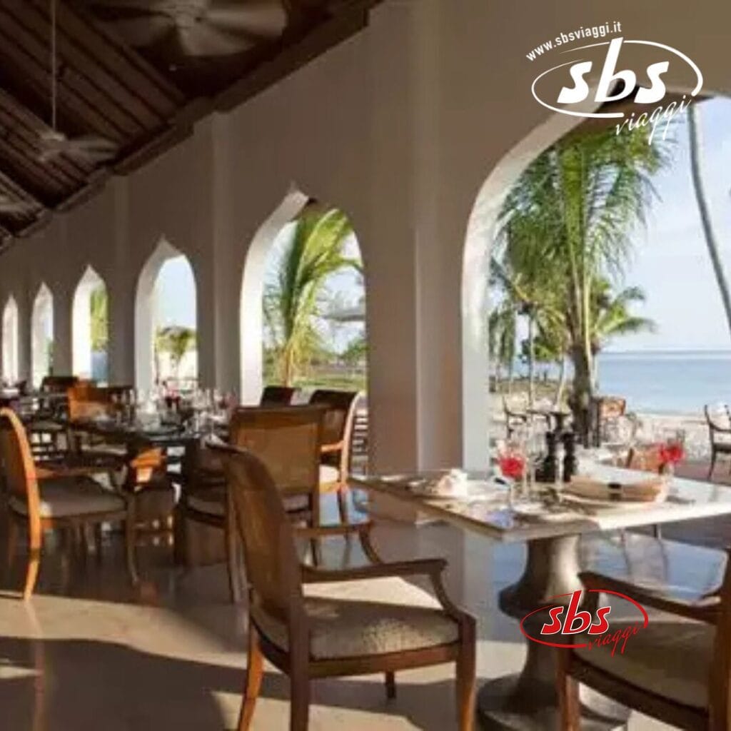 Un ristorante all'aperto presso The Residence, con tavoli e sedie in legno, finestre ad arco e una vista mozzafiato sulle palme e sull'oceano di Zanzibar.