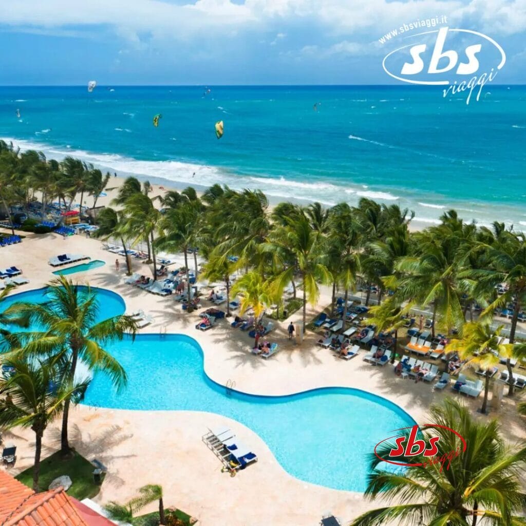 Resort sulla spiaggia con una grande piscina circondata da palme, con vista sull'oceano. La gente si rilassa mentre i kitesurfer danzano sulle onde, aggiungendo eccitazione a questa cornice idilliaca. Il logo Bozza Automatica adorna l'angolo in alto a destra, catturando l'essenza del relax e dell'avventura.