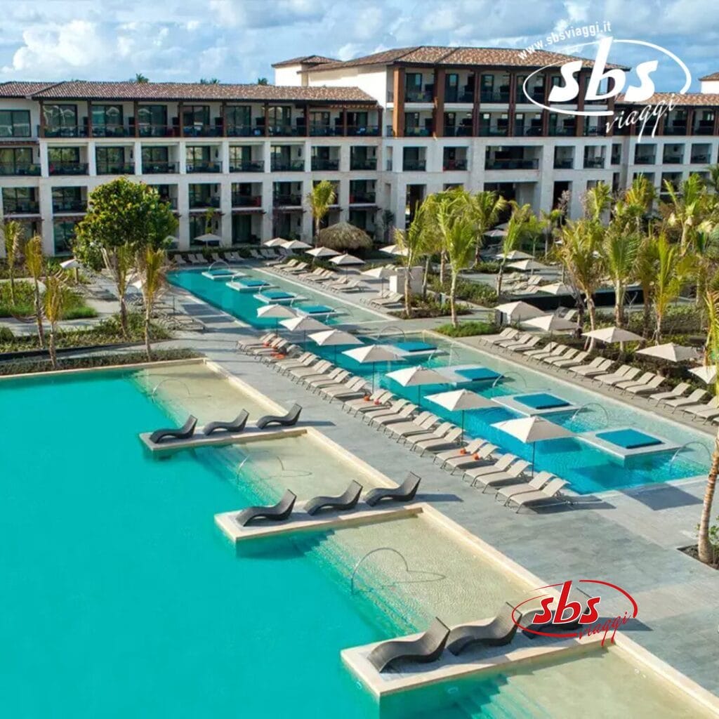 Vista aerea di un resort con più piscine, sedie a sdraio e palme che ondeggiano nella brezza. Un edificio a più piani si erge maestoso sullo sfondo, offrendo un perfetto mix di lusso e relax, quasi come se fosse stato catturato da Bozza automatica.