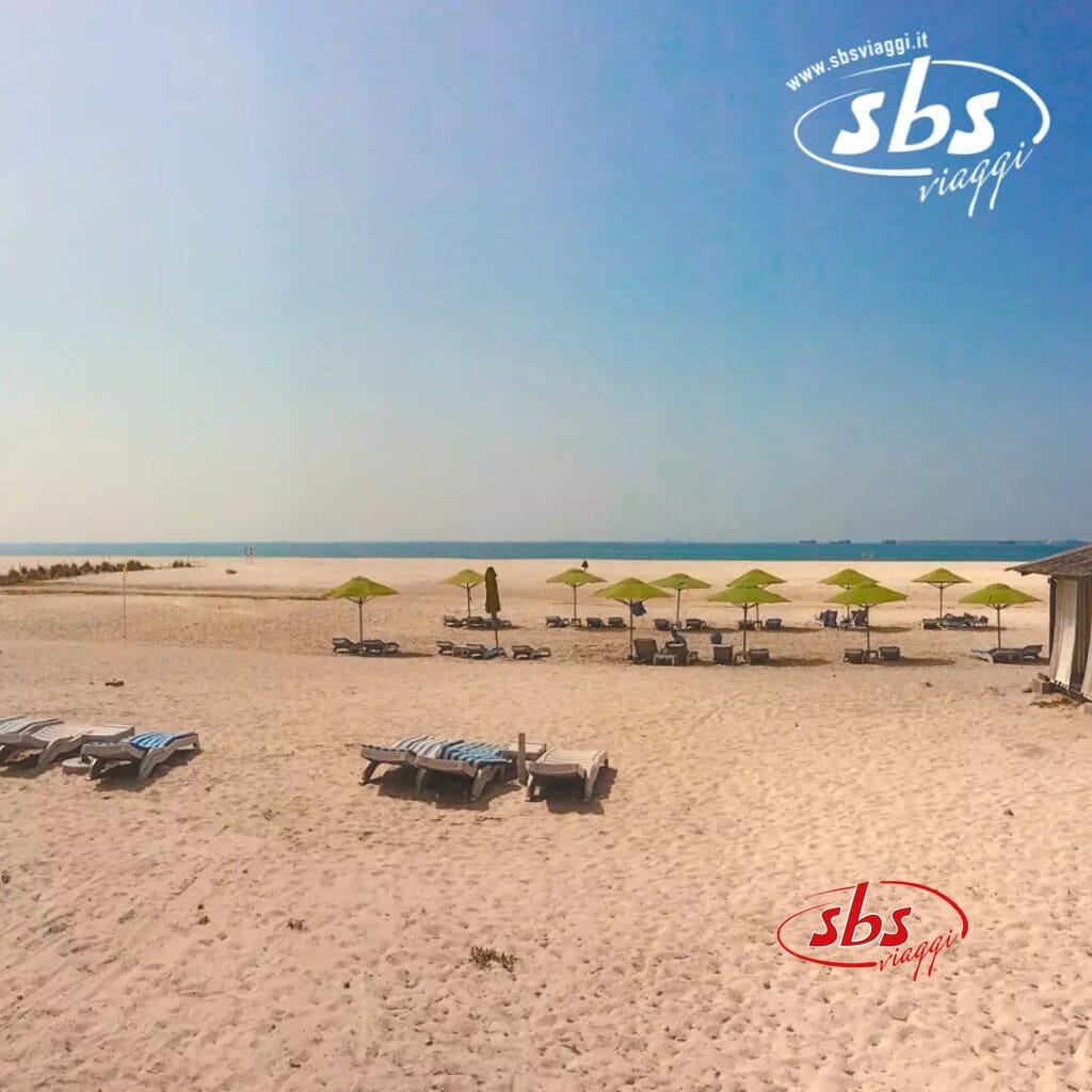 La tranquilla spiaggia sabbiosa dell'Hilton Resort di Salalah è attrezzata con numerose sedie a sdraio e ombrelloni verdi, tutti perfettamente allineati verso l'oceano sotto un cielo azzurro e limpido.