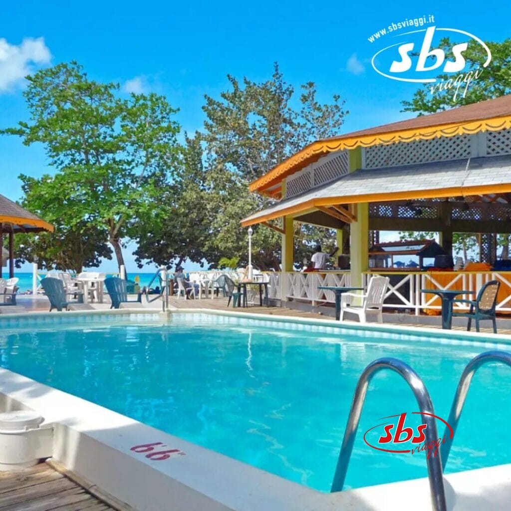 La piscina esterna del Merril's Beach Resort è dotata di una scala di metallo, circondata da tavoli e sedie. Alberi e un edificio con un tetto giallo esaltano lo scenario, mentre il mare blu luccica in lontananza.