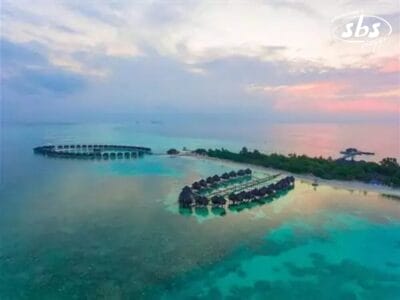 Vista aerea di bungalow sull'acqua e di un resort su una piccola isola, Bozza automatica, che cattura la serena bellezza delle acque turchesi dell'oceano sotto un cielo color pastello al tramonto.