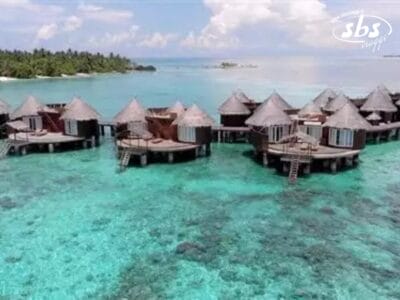 Bungalow su palafitte immersi in acque limpide e turchesi, con una vista in lontananza su una piccola isola e palme sotto un cielo parzialmente nuvoloso, creano uno scenario idilliaco come una composizione di Bozza.