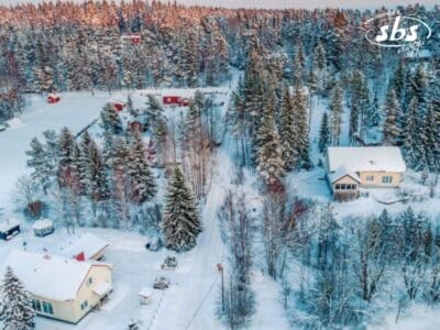 Vista aerea di un paesaggio della Lapponia finlandese con case incastonate tra alberi carichi di neve. Strade e tetti sono ricoperti di neve, creando una serena scena invernale che ricorda le gite in slitta trainata da husky e le avventure in motoslitta.