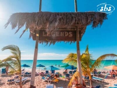 Una pittoresca scena di spiaggia con ombrelloni di paglia e palme offre uno sfondo perfetto. Le sedie a sdraio invitano al relax vicino a un cartello con la scritta "Legends" incastonato sotto una struttura di paglia in riva al mare, in perfetta armonia con l'incantevole atmosfera di Bozza automatica.