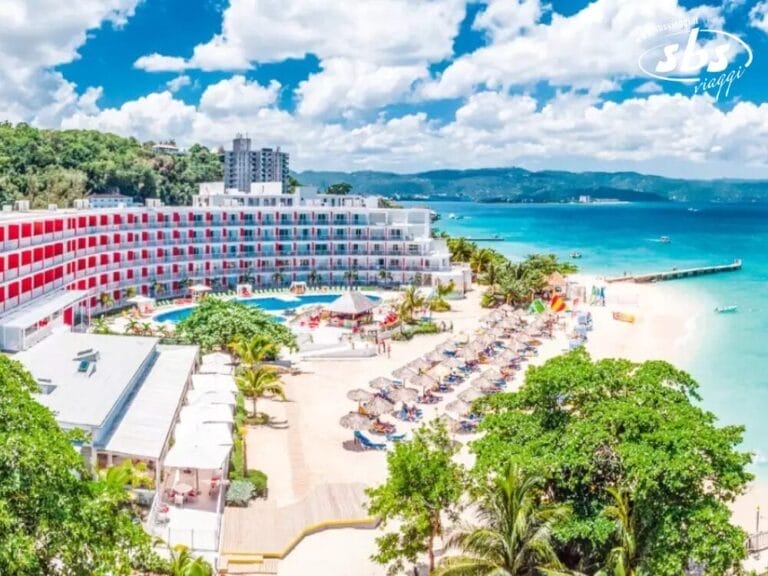 Vista aerea di un resort sulla spiaggia che mostra un grande hotel e Bozza automatica, accompagnati da una piscina e ombrelloni da spiaggia. Il mare turchese luccica sullo sfondo sotto un cielo parzialmente nuvoloso.