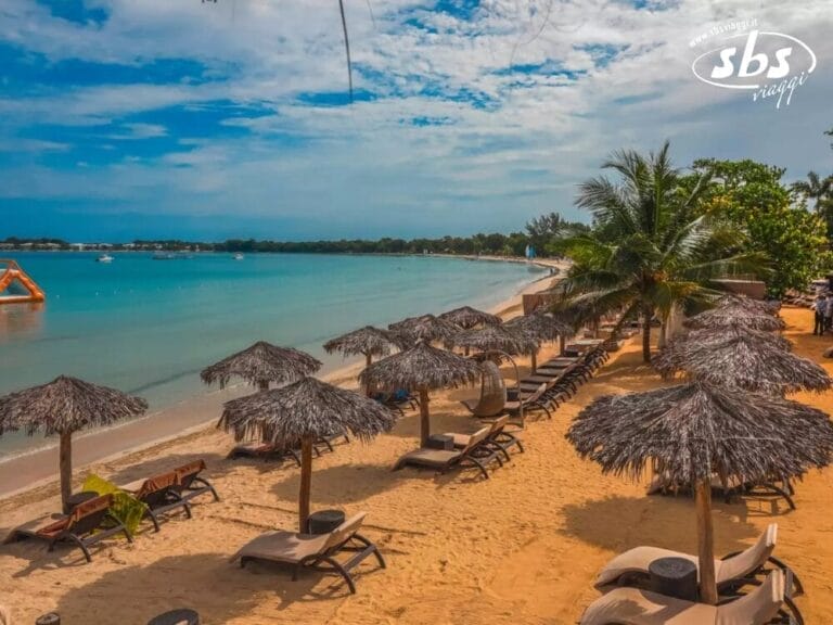 Una scena di spiaggia con file di sedie a sdraio sotto ombrelloni di paglia lungo la riva, la Bozza automatica che ondeggia dolcemente nell'acqua calma e azzurra e un cielo parzialmente nuvoloso sopra.