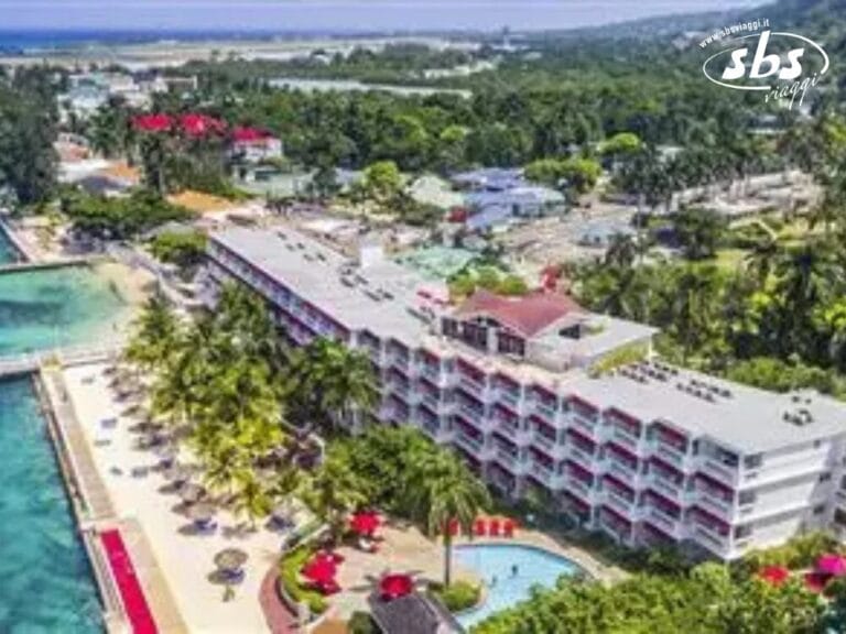 Vista aerea del resort sulla spiaggia Royal Decameron con diversi edifici, palme e ombrelloni rossi attorno a una piscina. Le sedie a sdraio invitano al relax a Montego Beach, con l'oceano e la vegetazione lussureggiante che offrono uno sfondo mozzafiato.