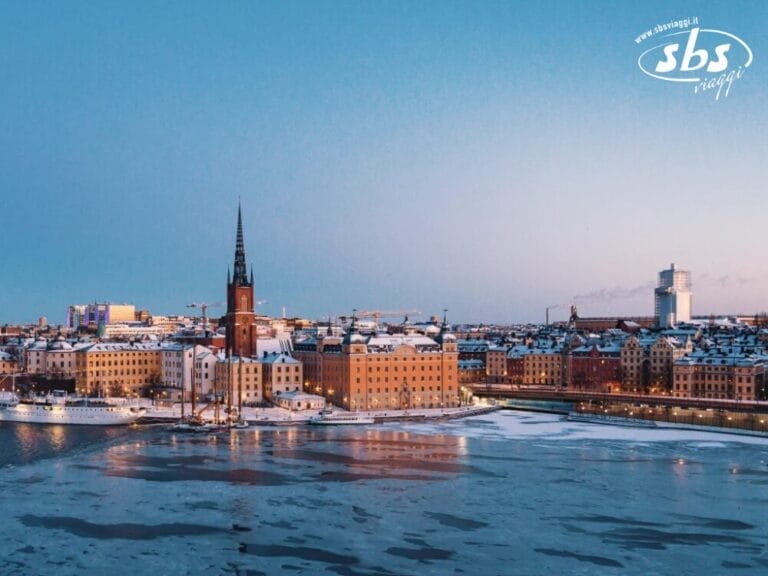 Un paesaggio urbano invernale cattura la magia delle capitali del Nord, mostrando un'area urbana coperta di neve con edifici e il campanile di una chiesa in riva a uno specchio d'acqua parzialmente ghiacciato sotto un cielo azzurro e limpido.